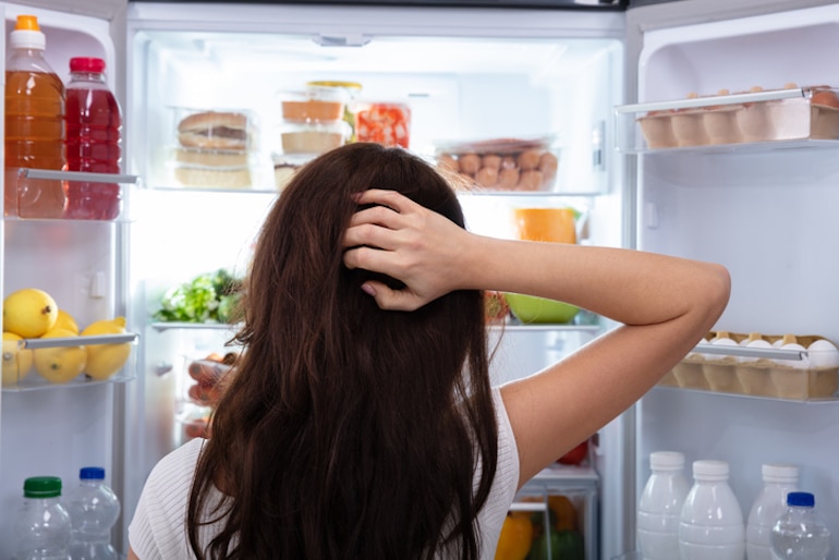 从后面拍摄的黑发女人看着冰箱，不知道在她的间歇性禁食打开吃饭窗口吃什么